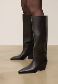 Svarta knähöga stövlar i läder med spetsig tå och klack med blockhäl, med en slät yta och rena linjer.