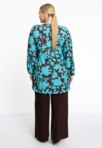 Tunique à manches longues avec un motif floral turquoise sur fond marron foncé, associée à un pantalon large marron. Tissu lisse, coupe décontractée.