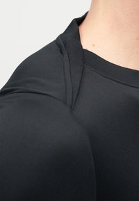 Κοντινή λήψη ενός ατόμου που φορά μαύρο πουκάμισο, εμφανίζοντας τη ραφή του ώμου και τις λεπτομέρειες του λαιμού σε ανοιχτόχρωμο δέρμα.