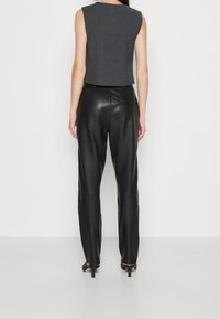 Frau trägt ärmelloses graues Oberteil und hoch taillierte schwarze Lederhose, steht mit dem Rücken zur Kamera vor einem schlichten weißen Hintergrund.