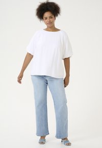 Weiße, oversized Baumwollbluse mit kurzen Puffärmeln, kombiniert mit hellblauen Jeans. Die Schuhe sind blaue High Heels mit ausgefransten Riemen.