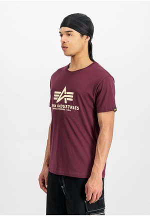 Bordeaux bomulds t-shirt med en cremefarvet logo "Alpha Industries" og detaljer; korte ærmer og rund halsudskæring, båret med sorte bukser.