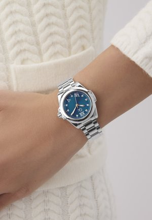 Srebrny metalowy zegarek z niebieską tarczą, rzymskimi cyframi i diamentowymi znacznikami godzin, oraz ogniwowym paskiem. W tle widoczny dziany kremowy sweter.