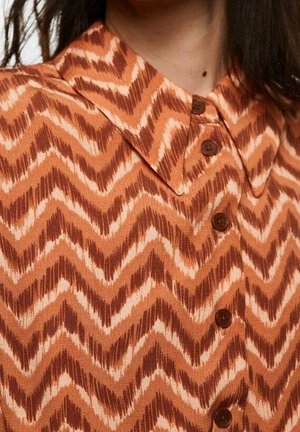 Overhemd met kraag en een zigzagpatroon in bruin, beige en oranje, met bruine knopen aan de voorkant, gedragen door een persoon met donker haar.