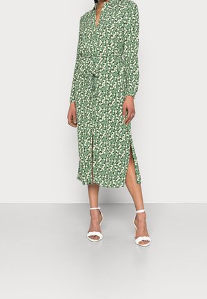 Femme portant une robe midi verte à motifs floraux avec manches longues, fentes devant et sur les côtés, et des sandales blanches à talons hauts avec brides à la cheville.