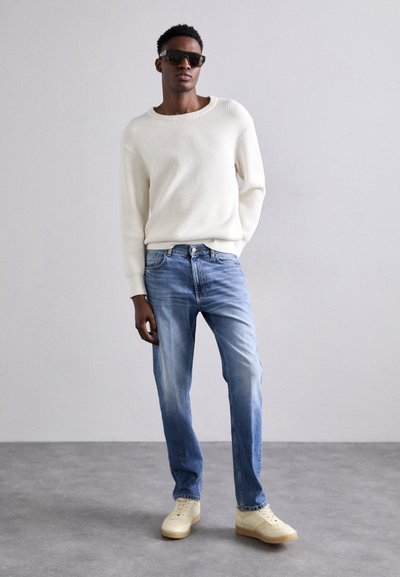 Fehér bordázott pulóver, világoskék farmer és krém színű sportcipő. Modell sötét napszemüveget visel, és egy világos szürke háttér előtt áll.