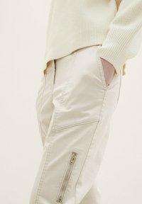 Personne portant un pantalon beige clair avec une poche zippée latérale et un pull côtelé crème, main partiellement dans la poche, fond neutre.