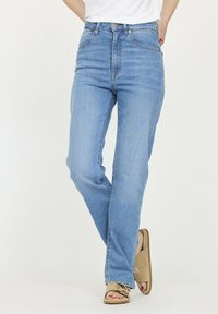 Personne portant un jean taille haute bleu clair et des sandales en suède ouvertes tannées, se tenant avec une jambe croisée devant l'autre.