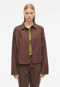 Bruine button-up jas met een gestructureerde kraag, oversized mouwen en een textuurafwerking. Gedragen over een groene top en gecombineerd met een bruine broek.