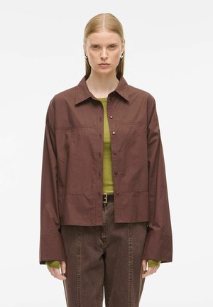 Brown button-up jacket with a structured collar, oversized sleeves, and a textured finish. Worn over a green top and paired with brown pants.