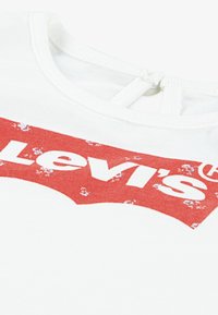 Fehér pamut póló piros logó grafikával, amelyen a "Levi's" fehér színnel szerepel. Kerek nyakkivágás, hátul gomblyuknyílással.
