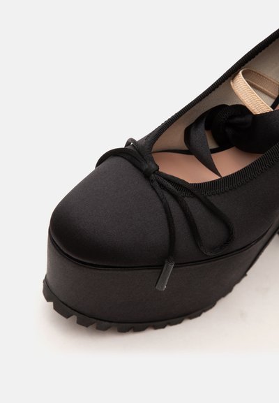 Chaussure à plateforme noire avec semelle épaisse, bout arrondi, bride élastique et nœud décoratif à l'avant.