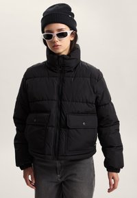 Giacca a vento nera con collo alto, chiusura con cerniera e due tasche frontali con bottoni a pressione. Indossata con un berretto nero e occhiali bianchi.