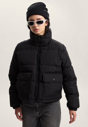 Doudoune noire avec col montant, fermeture éclair et deux poches avant à boutons-pression. Portée avec un bonnet noir et des lunettes blanches.