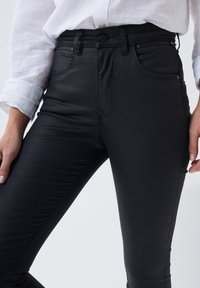 Jeans ajustados de mezclilla negra con una cintura alta, bolsillo con cremallera en la parte delantera y detalles de costura sutiles. El material tiene un acabado suave y ligeramente brillante.