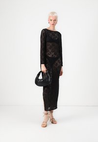 Vestido preto em renda com mangas longas, silhueta ajustada, fendas laterais e uma bolsa preta a condizer. A modelo usa sandálias de tiras beijas.