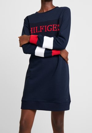 Personne portant une robe bleu marine avec des poignets rayés rouges et blancs et un texte rouge "HILFIGER" sur la poitrine, les bras croisés.