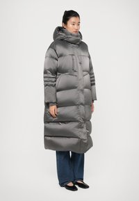 Lange, puffy grijze pufferjas met een hoge kraag, ritsclosure en doorstikte patroon. Draagt over blauwe wijde jeans en zwarte schoenen.