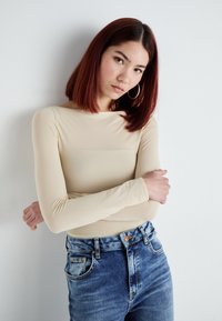 Beige hosszú ujjú felső, sima, testhezálló kialakítással, magasan elhelyezett kék farmernadrággal párosítva, amely elhasznált textúrával és klasszikus gombos záródással rendelkezik.