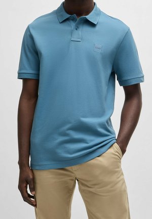 Mand iført en lyseblå kortærmet polo shirt med lille logo på brystet og beige bukser, den ene hånd i lommen.