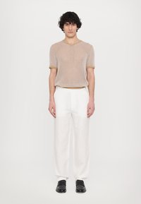 Top en tricot beige clair à manches courtes avec une texture côtelée et des finitions dorées contrastantes, associé à un pantalon en lin blanc ample et des chaussures noires.