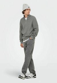 Grijze trui met rits en lange mouwen, gedragen over een wit overhemd, gecombineerd met grijze spijkerbroek en zwart-witte sneakers en een lichtgrijze muts.