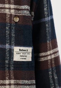 Camisa de flanela em xadrez azul escuro, castanho e branco. Tecido texturizado com um rótulo creme que apresenta detalhes da marca e do produto. Fecho com um único botão.