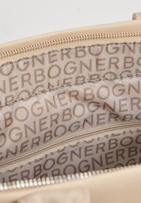 Notranjost bež torbe z ponavljajočim se vzorcem besedila "BOGNER", ki prikazuje žep na zadrgo in vidne šive ob oblogi.