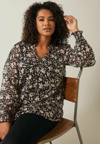 Blusa floreale nera e crema con maniche lunghe a sbuffo, tessuto leggero e trasparente, e colletto con bottoni; indossata sopra pantaloni neri aderenti.