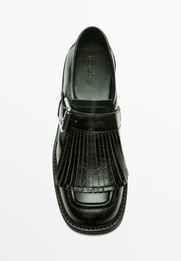 Sort lædersko med fladt design, der har en frynset front, rem med spænde og syet detalje langs sålen.