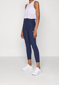 Lichtpaars mesh crop top gecombineerd met marineblauwe high-waisted leggings. De leggings hebben een zijvak en een subtiel logo-detail. Witte sportschoenen.