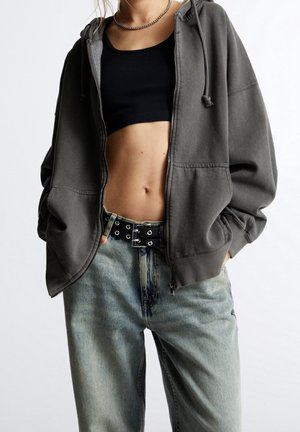 Personne portant un sweat à capuche gris foncé oversize avec fermeture éclair, un top court noir, un jean bleu clair avec ceinture noire, debout les mains dans les poches.