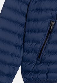 Acercamiento de la manga y lado de una chaqueta acolchada azul marino con un bolsillo negro con cremallera y dobladillo elástico.