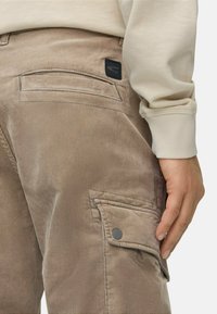 Beige corduroy cargo bukser med sidelommer, baglomme med syning og logo patch. Hånd hviler på lommen.