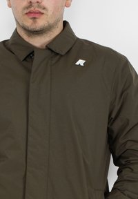 Giacca leggera verde oliva con colletto alto, caratterizzata da una discreta toppa con logo sul petto e una superficie liscia.