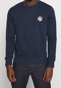 Man som bär en marinblå sweatshirt med en liten hexagonal vit och orange logotyp på vänster bröst, i kombination med mörkblå jeans.