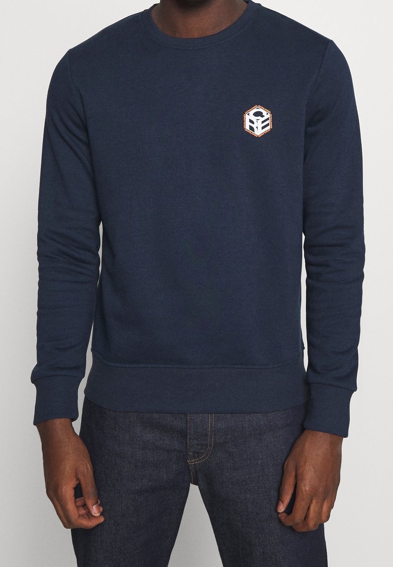 Man som bär en marinblå sweatshirt med en liten hexagonal vit och orange logotyp på vänster bröst, i kombination med mörkblå jeans.