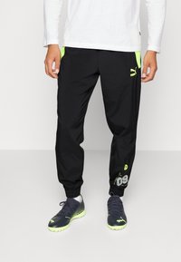Schwarze sportliche Hosen mit neongrünen Akzenten, elastischen Bündchen und Logodetails. Kombiniert mit schwarzen Sneakers, die grüne Sohlen haben.