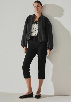 Moteris stovi prie sienos apsirengusi juodą bomber tipo striukę, baltą palaidinę su taškeliais ir nėriniais, juodus trumpintus kelnes ir juodus butus.