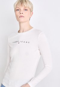 Svijetlo bež majica dugih rukava s okruglim izrezom, s crnim logotipom "TOMMY JEANS" na prsima. Meka, glatka tekstura tkanine.
