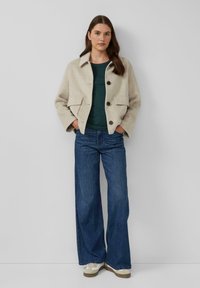 Vrouw in een beige knoopjasje, donkergroene shirt, wijde blauwe jeans en witte sneakers, staande tegen een effen lichte muur.