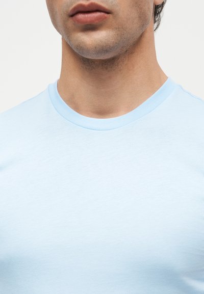 Κοντινό πλάνο στο κάτω μέρος προσώπου και λαιμό άνδρα που φοράει ανοιχτό μπλε κοντομάνικη μπλούζα με στρογγυλή λαιμόκοψη, μπροστά από απλό φόντο.