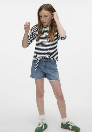 Maglietta a righe con maniche lunghe legata in vita, abbinata a pantaloni shorts di jeans. Sneakers verdi con lacci bianchi e calzini alla caviglia bianchi con volant.