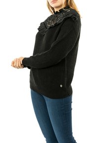 Maglione nero lavorato a maglia con una finitura testurizzata, caratterizzato da un colletto in pizzo arricciato con decorazioni e un piccolo logo sul lato inferiore.