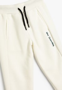 Crèmekleurige joggers met ribstructuur, zwarte trekkoorden en geborduurde "New Generation" detail aan de linkerkant.