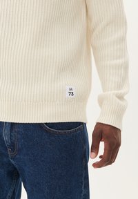 Grietos tekstūros, šviesiai kreminės spalvos megztinis su ilgomis rankovėmis, pagamintas iš medvilnės, su etikete "M 73", siuvinėta apačioje. Derinamas su tamsiai mėlynomis džinsomis.