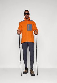 Oranž fleece-jope sinise taskuga, mustad retuusid ja matkakepid. Kangal on struktuurne välimus; kannab päikeseprille ja matkasaapaid.