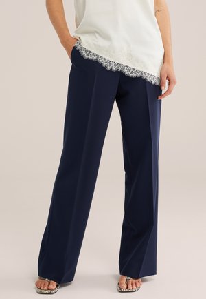 Mujer con pantalones anchos azul marino, top blanco con encaje y sandalias de estampado de piel de serpiente con punta abierta, de pie con las manos en las caderas.