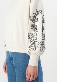Persona che indossa una maglietta bianca a maniche lunghe con ricami astratti neri sui polsini e jeans di denim blu, mostrata dalla vita alla mano.