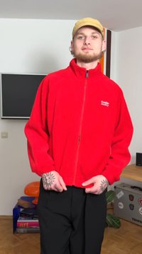 Piros polár cipzáras dzseki magas gallérral és elasztikus mandzsettákkal, fekete nadrággal párosítva. Bal mellrészen logóval díszítve.